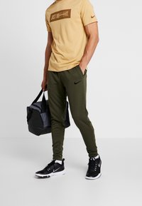 Ljusgul t-shirt med brun text, olivgröna joggers med en svart Nike-logotyp, svarta träningsskor och en svart gymväska med grå accenter.