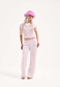 Világos rózsaszín kétdarabos összeállítás, amely egy rövid felsőből "JUICY Angel" felirattal és hozzáillő, megkötős nadrágból áll. A modell bolyhos rózsaszín sapkát visel.