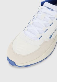 White and blue sneaker featuring a mix of smooth fabric and suede, with textured laces and a blue rubber sole. Brand logo is visible.