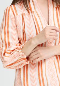 Personne ajustant le poignet d'une blouse à manches longues avec des rayures verticales orange, rose et blanche et un motif texturé.