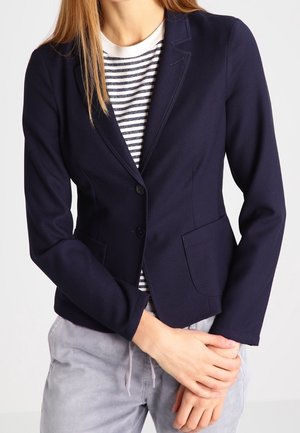 Kvinde iført en tætsiddende navy blazer over en sort-hvid stribet skjorte med lysegrå bukser, stående mod en ensfarvet baggrund.