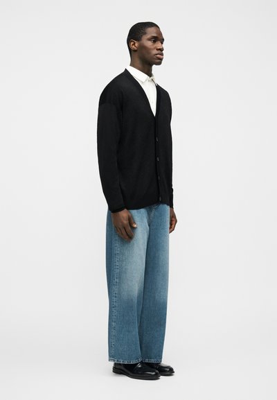 Ά νδρας που φορά μαύρη ζακέτα με κουμπιά, λευκό πουκάμισο με γιακά, φαρδιές ανοιχτόχρωμες μπλε τζιν και μαύρα παπούτσια, σε απλό φόντο.