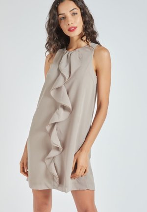 Mujer con un vestido sin mangas, beige y de largo hasta la rodilla, con un volante vertical en la parte delantera, posando contra un fondo claro y liso.