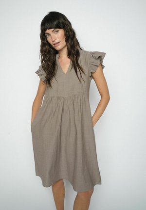 Kobieta z długimi, falującymi włosami, ubrana w luźną, beżową sukienkę sięgającą kolan, z krótkimi falbaniastymi rękawami, stojąca z rękami w kieszeniach na jednolitym tle.