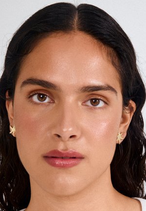 Woman with dark wavy hair, brown eyes, natural makeup, matte pink lips, and small gold star-shaped earrings, facing forward against a plain background.