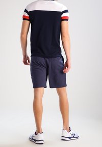 Marinblå pikétröja med kort ärm och vita och röda randiga detaljer, matchad med marinblå shorts och vita sportskor, sedd bakifrån.