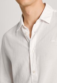 Camisa de lino blanca con cuello abotonado y pequeño logotipo bordado en el pecho, con botones visibles y una superficie de tela texturizada.