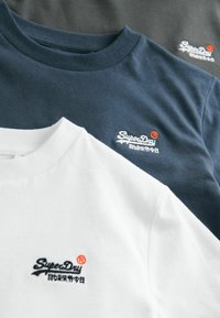 Tres camisetas apiladas: blanca, azul marino y negra. Cada una presenta un cuello redondo, material de algodón suave y un logo bordado "SuperDry" en colores contrastantes.