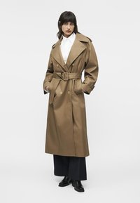 Femeie purtând un trench maro lung cu curea peste o cămașă albă, pantaloni negri largi și ghete negre până la gleznă, stând cu mâinile în buzunare.