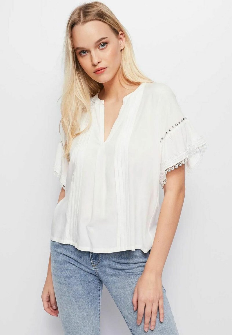 Blusa bianca con scollo a V, maniche corte a volant con bordo in pizzo e dettagli argentati. Realizzata in tessuto leggero con pieghe verticali.