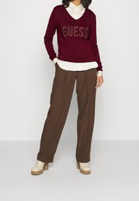 Bordeauxfarbener V-Ausschnitt-Pullover mit metallic "GUESS"-Logo, über einem weißen Hemd mit Kragen getragen, kombiniert mit braunen, weit geschnittenen Hosen und beigen Plateauschuhen.