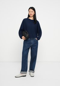 Tengerészkék, laza fazonú pulóver, amelyet bő szárú kék farmernadrággal és világosszürke slip-on cipővel visel. Fekete klutcs táska a kezében.