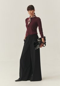 Bordeaux-farbene Langarmbluse kombiniert mit hoch taillierten, weit geschnittenen schwarzen Hosen. Das Outfit wird durch eine elegante schwarze Handtasche mit gebundenem Schal ergänzt.