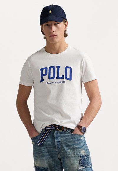 Biała bawełniana koszulka z niebieskim napisem "POLO" oraz tekstem "RALPH LAUREN". Noszona z przetartymi niebieskimi dżinsami i paskiem w paski. Klasyczna granatowa czapka.