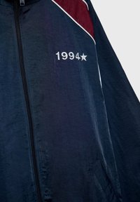Námořnická větrovka s vínovými ramenními akcenty, bílým lemováním a vyšívaným "1994★" na levém prsu. Obsahuje přední zip a boční kapsy.