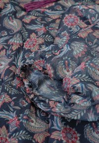 Floral patroon stof in donkerblauw, met rode en groene motieven. Lichtgewicht met een geplooide textuur vlakbij de rand. Aangedikt met franjes.