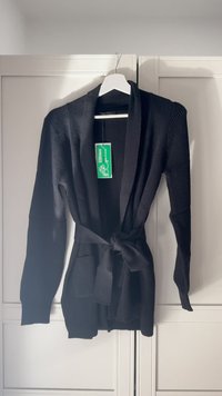 Schwarze Strickjacke mit strukturiertem Muster, langen Ärmeln, tailliertem Gürtel und Vordertaschen. Hängt an einem weißen Kleiderbügel vor hellem Hintergrund.