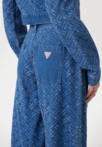 Blå denimjumpers med ett mönstrat tyg, med en bakficka med en röd och vit triangellogo. Långärmad och med oinskränkt midja.