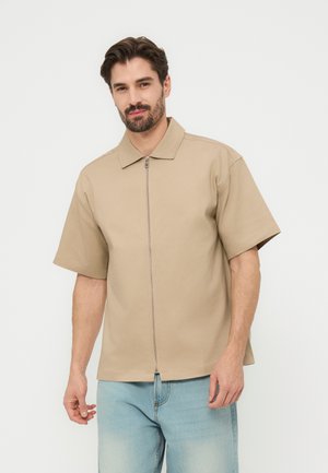 Mann mit dunklem Haar und Bart, der ein beiges kurzärmliges Zip-up-Hemd und hellblaue Jeans trägt, steht vor einem schlichten weißen Hintergrund.