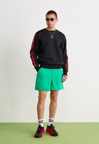 Hanorac negru cu dungi roșii pe laterale, pantaloni scurți verzi, șosete în dungi și adidași negri/roșii. Accesoriile includ ochelari de soare și un colier.