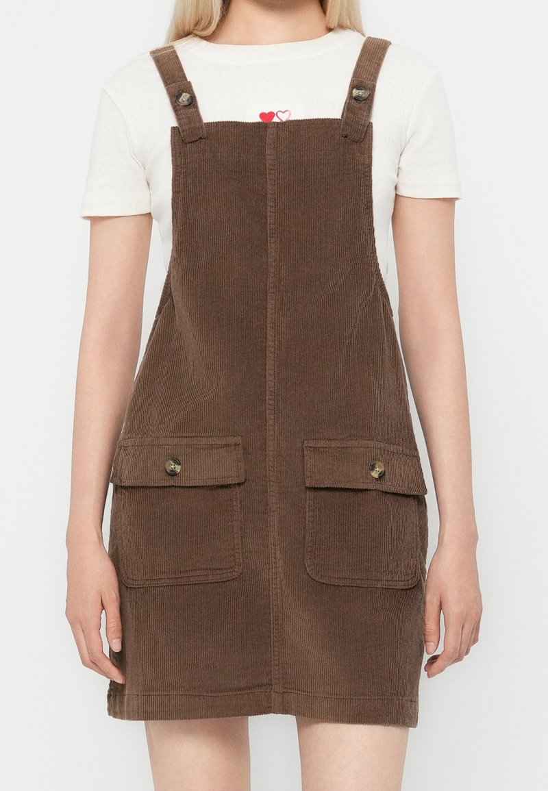 Femme portant une robe salopette en velours côtelé marron avec des poches avant boutonnées sur une chemise blanche à manches courtes avec un petit motif en forme de cœur.