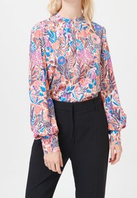 Blouse fleurie aux couleurs pastel avec manches longues bouffantes, col haut et texture douce et lisse. Portée avec un pantalon noir.