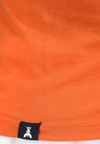 Oranžové bavlněné tričko se hladkým povrchem, se černou látkovou nášivkou na spodní straně s logem. Jednoduchý design, bez vzorů nebo akcentů.