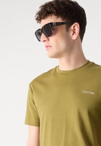 Tricou oliv verde cu mânecă scurtă, având logo-ul alb "Calvin Klein", asortat cu ochelari dreptunghiulari negri cu accente aurii.