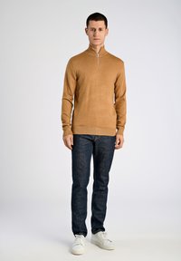Maglione zip in tessuto knit color marrone chiaro, abbinato a jeans blu scuro e sneakers bianche. Design semplice con polsini e orlo a costine.