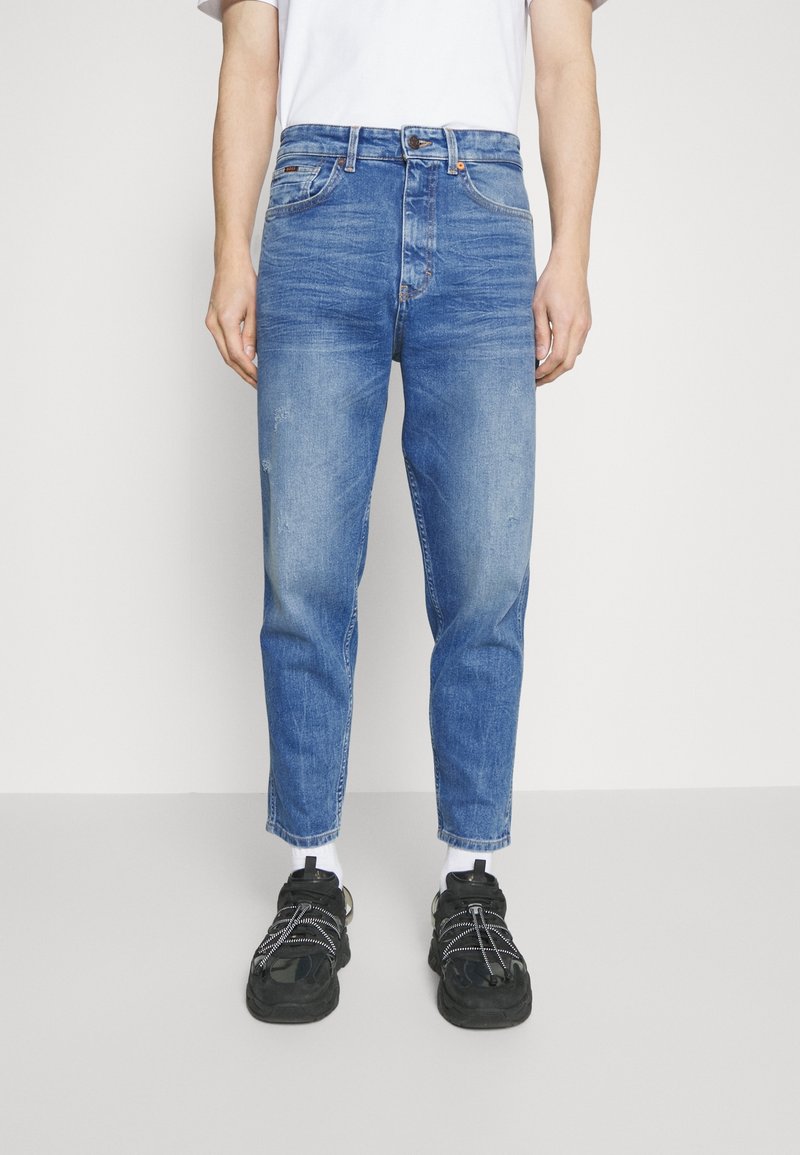 Siniset farkut, joissa on löysä malli, hieman haalistuneita, etutaskut ja vetoketjukiinnitys. Yhdistetty mustiin lenkkarikenkiin ja valkoisiin sukkiin.