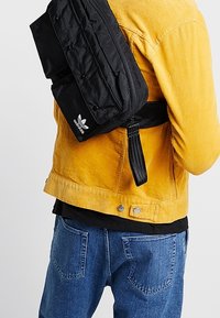 Person som bär en gul jacka och blå jeans med en svart Adidas axelväska över ryggen.