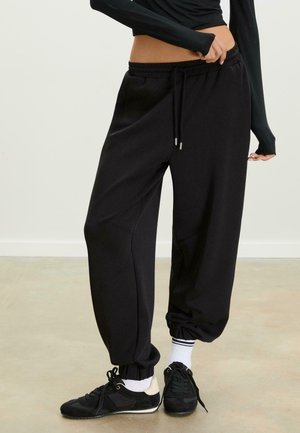Black sweatpants with an elastic waistband and drawstring. Tapered cuffs. Soft material with a smooth texture. Paired with black athletic shoes.
