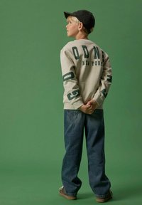 Grauer Sweatshirt mit grüner Aufschrift "OD NY", kombiniert mit lockeren blauen Jeans. Das Kind trägt eine schwarze Kappe und braune Schuhe.