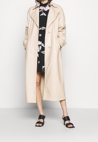 Beige, polvenpitkä trench-takki kiiltävästä materiaalista, jossa on olkaremmityyppiset olkatoppaukset ja suuret kultaiset napit, käytetty mustan kukallisen mekon päällä.