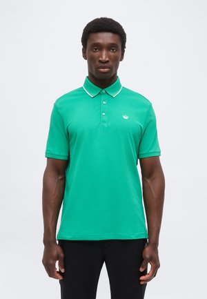 Man standing facing forward wearing a green short-sleeve polo shirt with white trim on the collar and black pants.
