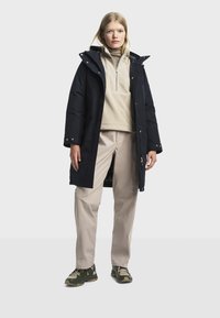 Femme portant un pull en polaire beige à col roulé, un pantalon beige clair, un manteau long noir à capuche et des chaussures de randonnée vert-beige, se tenant sur un fond blanc.