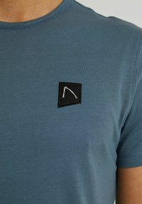 Modrá košile s krátkým rukávem, s malou černou čtvercovou nášivkou, na které je bílý abstraktní liniový design na horní části hrudi.