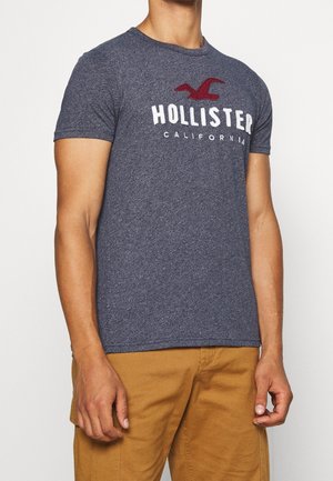 Mann trägt ein eng anliegendes navyblaues Hollister California T-Shirt mit einem roten Vogel-Logo, kombiniert mit hellbraunen Hosen.