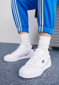 White sneakers with textured leather, a flat sole, and a black accent on the heel. Paired with blue track pants and white socks.