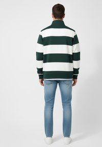 Hombre de pie de espaldas, con una camiseta de rayas horizontales verdes y blancas de manga larga, jeans azul claro y zapatillas blancas.