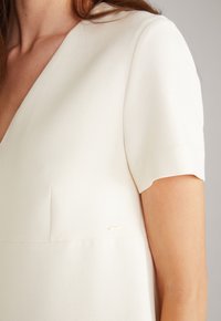Elfenbeinfarbiges Kleid mit V-Ausschnitt und kurzen Ärmeln. Glattes Material, ausgestattet mit einem dezenten Nahtdetail und einem kleinen goldenen Akzent.