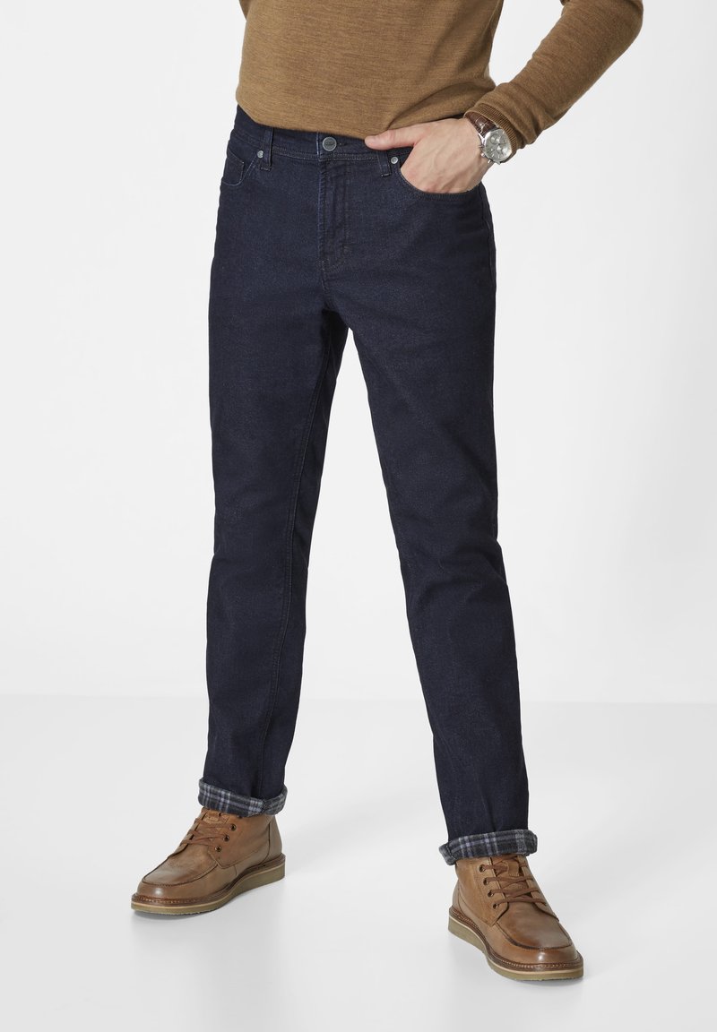 Dark blue denim jeans with a classic fit, five-pocket design, and cuffed hems. Worn with brown leather boots and a brown pullover.