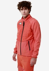 Giacca sportiva rossa con colletto alto, apertura con zip, dettagli neri e logo. Realizzata in tessuto liscio con design aderente e texture a contrasto.