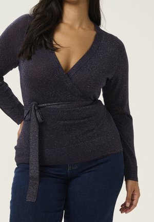 Woman wearing a dark sparkly wrap-style sweater with a tied belt and dark blue jeans, standing with one hand on her hip.