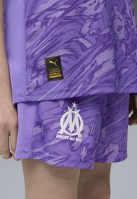 Pantaloni sportivi viola con un motivo testurizzato, logo ricamato e etichetta con il marchio. Il materiale sembra leggero, adatto per lo sport.