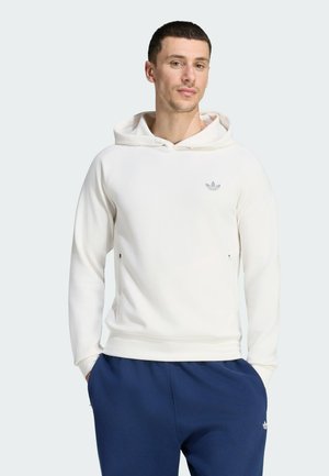 Mann mit kurzen Haaren, der einen weißen Kapuzenpullover und blaue Hosen trägt, steht mit den Händen in den Taschen vor einem einfarbigen Hintergrund.