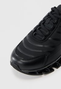 Svart atletisk sko med teksturert overdel med striper, rund tå og en polstret såle med tydelige gripselementer.