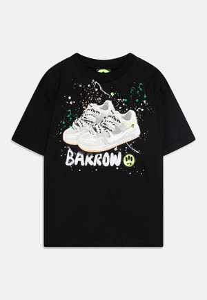 T-shirt noir avec un graphisme de baskets blanches avec des accents gris, des détails de lacets et un arrière-plan splatter peint en vert, blanc et marron.