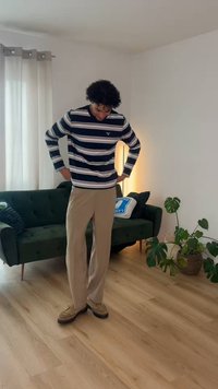 Άνδρας που φοράει μπεζ παντελόνι και ριγέ πουκάμισο με μακριά μανίκια στέκεται κοιτάζοντας προς τα κάτω σε ένα καθιστικό με πράσινο καναπέ και φυτά εσωτερικού χώρου.