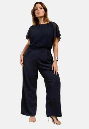 Frau trägt einen dunkelblauen Overall mit transparenten flatternden Ärmeln, weit geschnittene Hosen, silbernes Armband und beige hochhackige Sandalen, steht selbstbewusst.